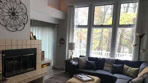 A living room with a fireplace, a sofa, and a coffee table. There are large windows with a view of trees. - Peaceful and Secluded! Lake Harmony Beautiful Townhome Ski Slope and Lake Views (Lake Harmony)