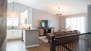 A modern kitchen with white cabinets, a central island, and a dining area. There is a living room with a brown sofa and a TV. - Centrally Located 2BR 2BA Home - Garage Parking & Fully Equipped Kitchen/Laundry (Janesville)