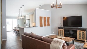 A modern living room with a brown leather sofa, a wooden coffee table, a flat-screen TV, and a kitchen area in the background. - Centrally Located 2BR 2BA Home - Garage Parking & Fully Equipped Kitchen/Laundry (Janesville)