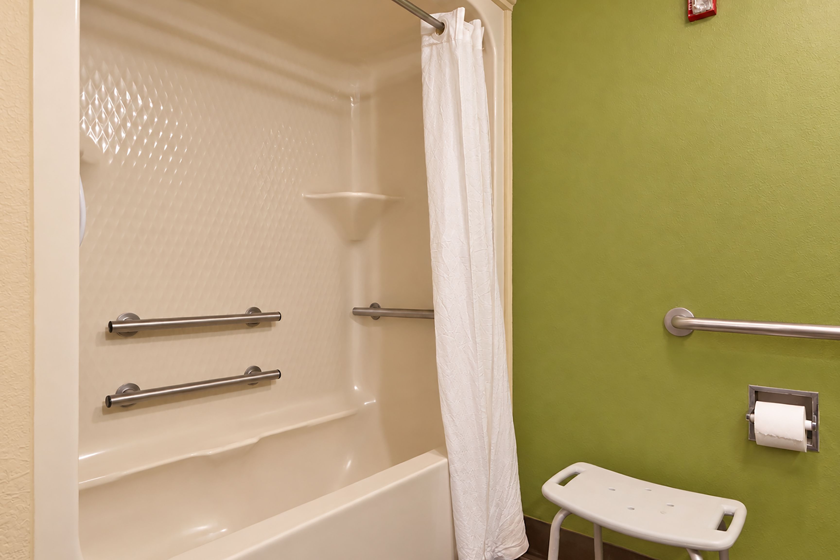 A bathroom with a bathtub, grab bars, a white shower curtain, a toilet paper holder, and a small white stool.