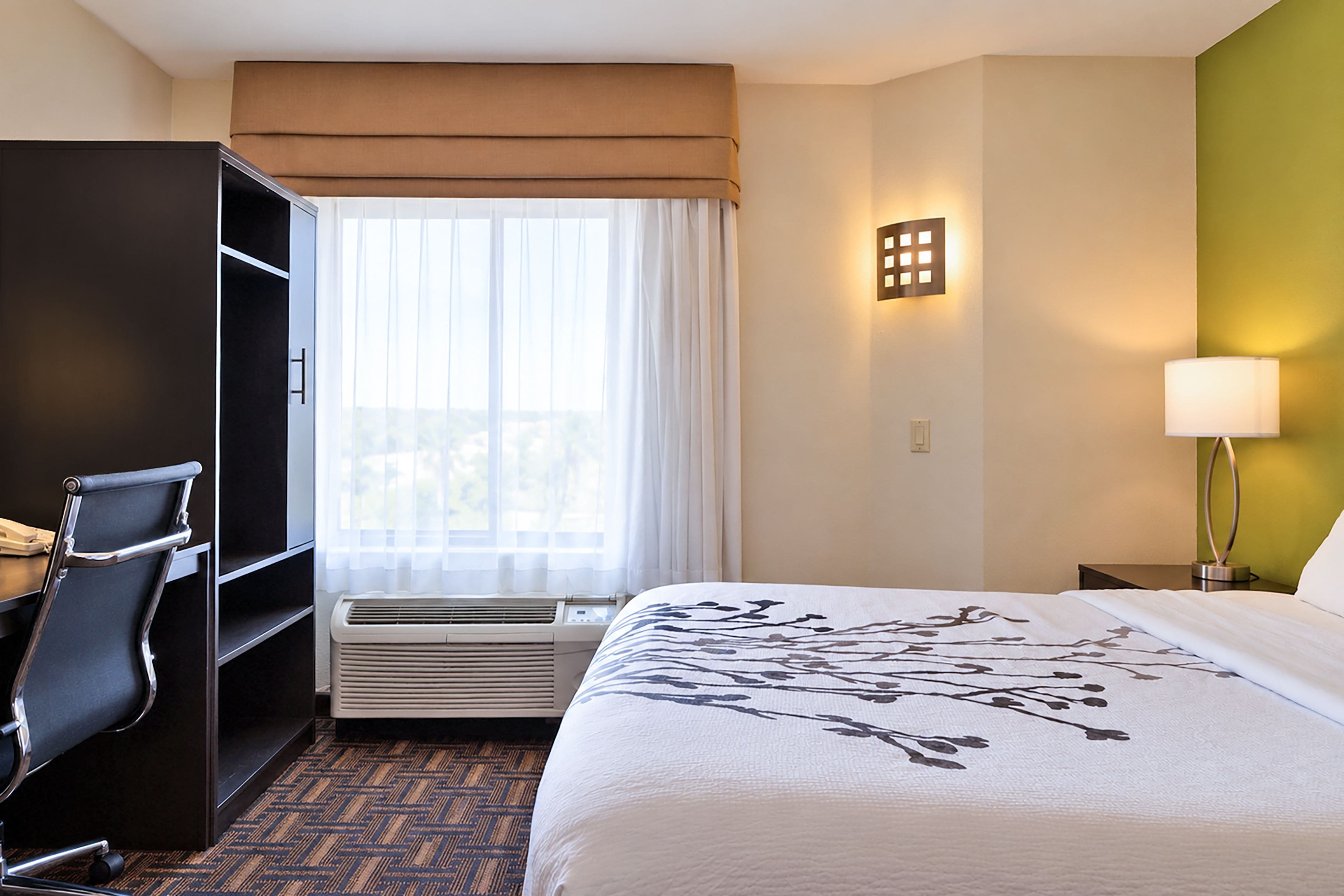 A hotel room with a bed, a desk, a chair, a wardrobe, a window with curtains, a wall-mounted light, and a wall heater.