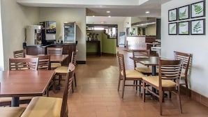 Un espace repas avec des tables et des chaises en bois, un coin cuisine équipé d’appareils électroménagers et des reproductions encadrées de gravures botaniques accrochées au mur.