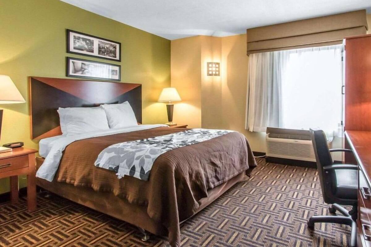 Een hotelkamer met een bed, twee bedlampen, een bureau met stoel, een raam met gordijnen en een wandairconditioner.
