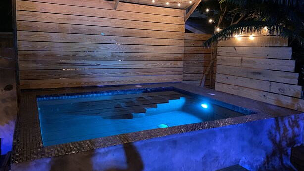 Een hottub met een houten terras en lichtslingers, omgeven door een houten omheining en palmbomen.