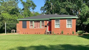 A single-story brick house with a green roof, a front door, and a small porch area. - The Edinburgh Cottage (Baton Rouge)