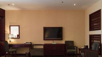 Habitación de hotel con televisor de pantalla plana montado en la pared, un escritorio con lámpara, dos sillones y un armario.