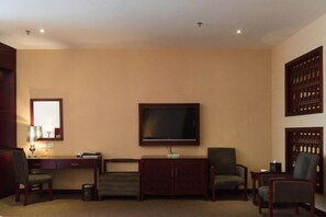 Habitación de hotel con televisor de pantalla plana montado en la pared, un escritorio con lámpara, dos sillones y un armario.