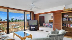 A spacious living room with a large TV, comfortable seating, a balcony with a view, and a bookshelf. - KAUAI BEST DEAL!!!!   1-10-25 thru 1-17-25    - Sleeps 8 (Koloa)