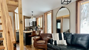 A cozy living room with a black leather sofa, wooden flooring, and a kitchen in the background. - Eagle Ridge Treehouse (Stockbridge)