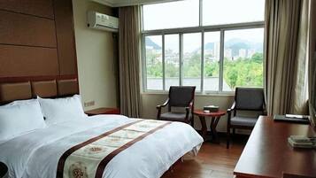 A hotel room with a large bed, two chairs, a small table, and a view of a cityscape through a large window.