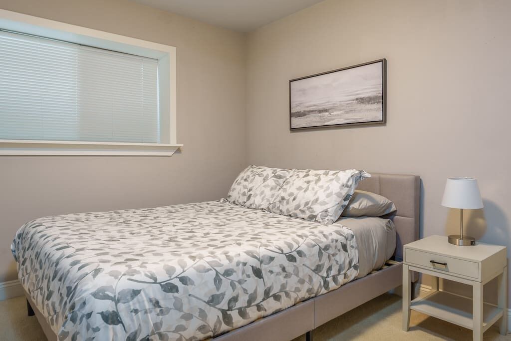 A bedroom with a bed, a nightstand, a lamp, a window with blinds, and a framed picture on the wall.
