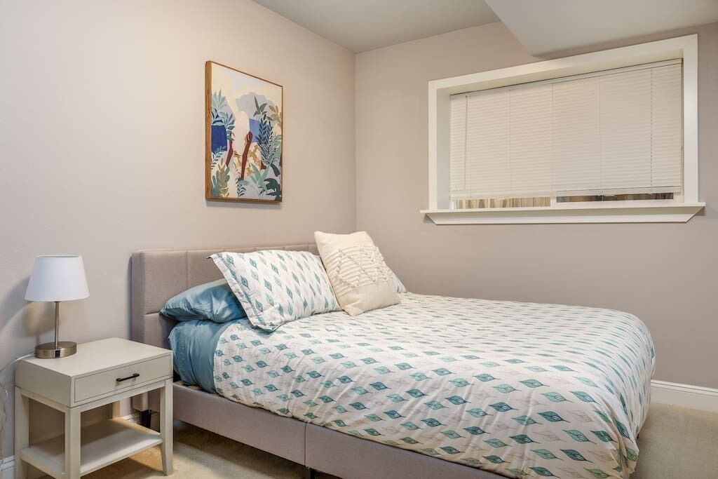 A bedroom with a bed, a nightstand, a lamp, a window with blinds, and a wall-mounted painting.
