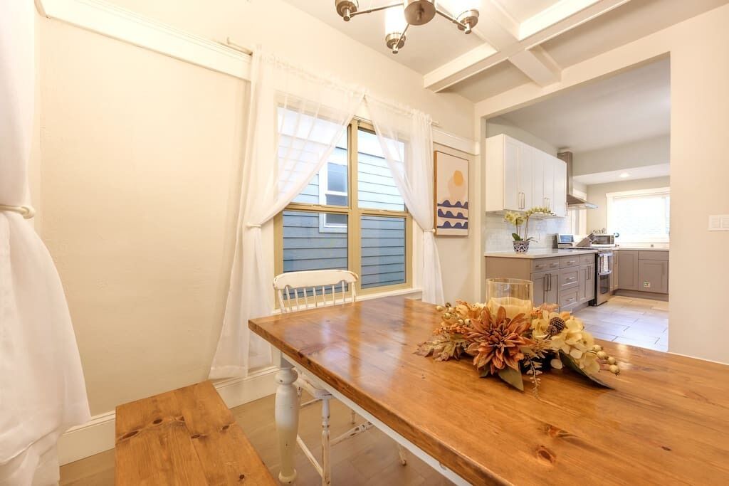A dining area with a wooden table, white curtains, and a view into the kitchen.