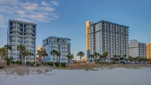Un hotel frente a la playa con varios edificios altos, palmeras y una playa de arena.