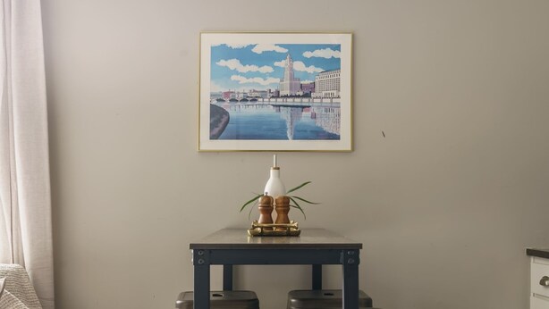 A dining area with a table and four stools, a framed cityscape picture on the wall, and a white cabinet to the right.
