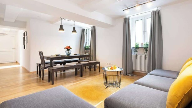 Ein modernes Wohnzimmer mit Essbereich, grauen Sofas, einem kleinen runden Tisch mit Obst und Blick ins Freie durch große Fenster.