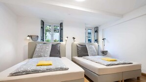 Zwei Einzelbetten mit grau-weißer Bettwäsche, ein kleiner Tisch und ein Fenster mit Blick auf Grünflächen.
