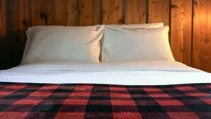 Una cama con sábanas blancas y una manta de cuadros frente a una pared de madera.