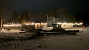 Un espacio exterior cubierto de nieve con un edificio pequeño, una zona delimitada por una valla con luces de cuerda y una silla roja.