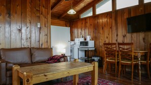 Un interior de cabaña acogedor, con piso de madera, un sofá de cuero marrón, una mesa de madera, una zona de cocina con microondas y cafetera, y un televisor montado en la pared.
