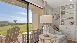 A living room with a view of a grassy area, a glass door leading outside, a white sofa, a small table with a lamp, and a white cabinet with shelves. - Capistrano E101. Gulf Front, Direct private beach access, Pet Friendly. (Seacrest)