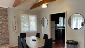 A dining area with a wooden table and chairs, a brick wall, a window with a view of trees, and a kitchen in the background.