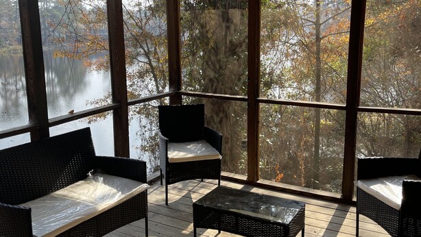 A covered outdoor seating area with wicker furniture and a glass table, overlooking a body of water and trees.