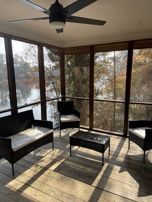 Un espace extérieur aménagé, abrité et doté de meubles en osier et d’une table en verre, offrant une vue sur un plan d’eau et des arbres.