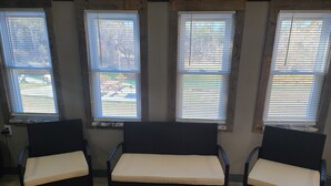 A room with three wicker chairs and a wooden bench, arranged in a semi-circle facing three windows with blinds. - Cabin 1 at the Pallet Factory (Leadwood)