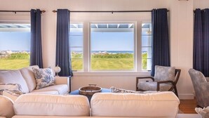 A room with a large window overlooking a grassy area and the sea. There is a white sofa and two chairs. - Miramar Escape (North Hampton)