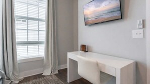 Ein Zimmer mit großem Fenster, einem flachbildfernseher an der Wand, einem weißen Schreibtisch und einem kleinen dekorativen Gegenstand auf dem Schreibtisch.