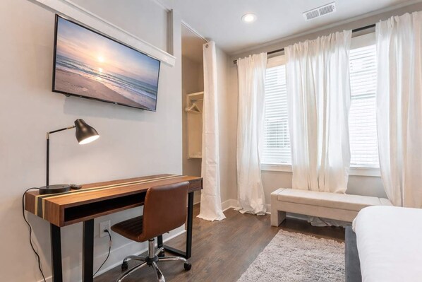 Habitación de hotel con una cama, un escritorio con silla, un televisor montado en la pared y una ventana con cortinas.