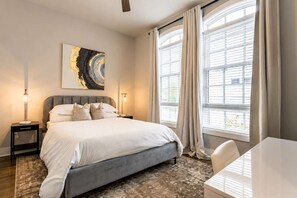 Ein modernes Schlafzimmer mit einem großen Bett, Nachttischlampen, einem Schreibtisch und Blick ins Freie durch große Fenster.