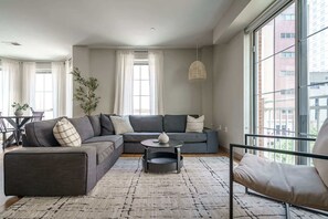 Uma sala de estar moderna com um sofá seccional cinza, uma mesa de centro redonda e uma janela grande com cortinas.