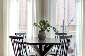 Mesa de jantar redonda com quatro cadeiras, vaso com folhagem verde e vista para uma varanda através de grandes janelas.