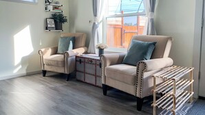 A living room with two beige armchairs, a small side table, a wooden shoe rack, and a window with curtains. - Washburne Studio: King size comfort and private Hot tub (Springfield)