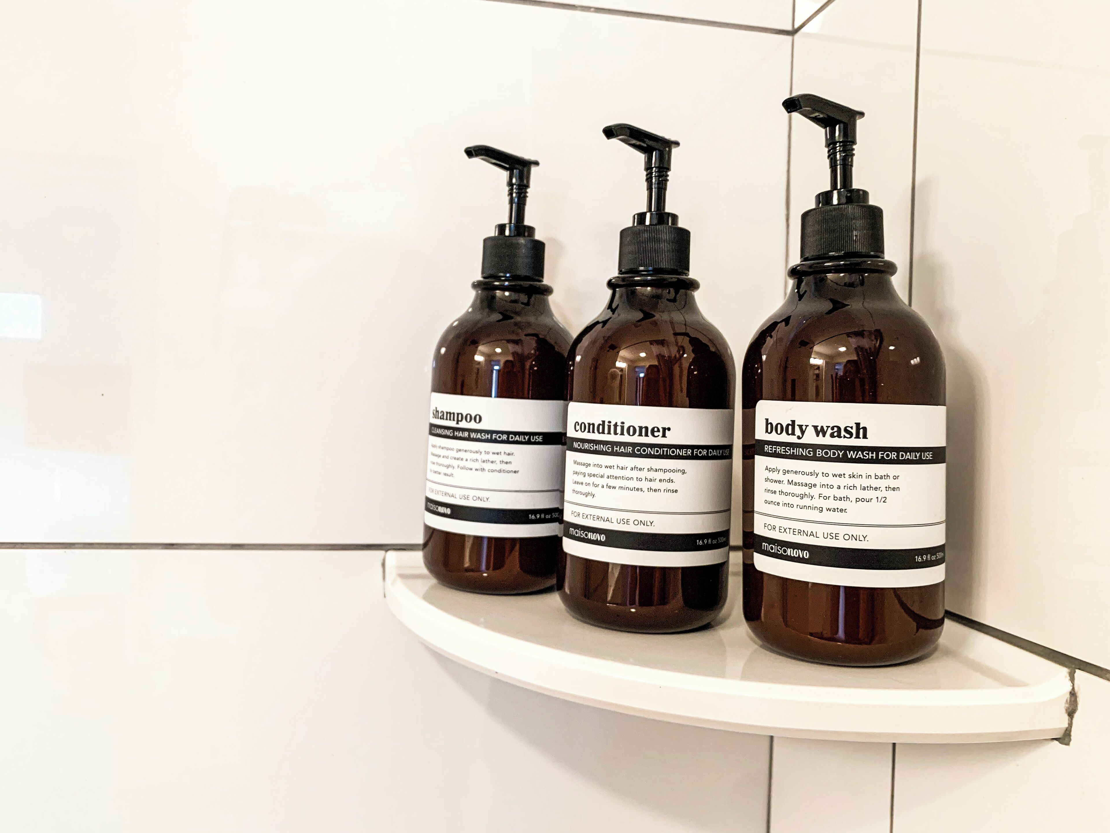 Drie flessen douchegel, conditioner en shampoo op een witte plank.