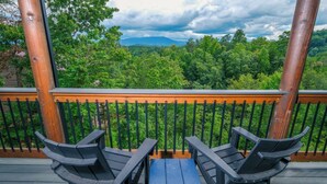 A wooden deck with two Adirondack chairs and a small table, overlooking a forested landscape with mountains in the distance. - Grace Place - Private Indoor Pool, Hot Tub, Views, Theater, and more! (Sevierville)