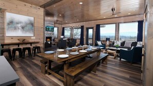 A spacious dining area with a long wooden table set for four, surrounded by comfortable seating and large windows offering a view. - Grace Place - Private Indoor Pool, Hot Tub, Views, Theater, and more! (Sevierville)