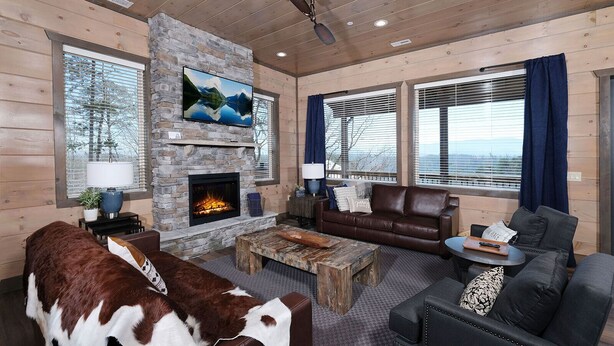 A cozy living room with a stone fireplace, leather furniture, and a large TV mounted on the wall. - Grace Place - Private Indoor Pool, Hot Tub, Views, Theater, and more! (Sevierville)