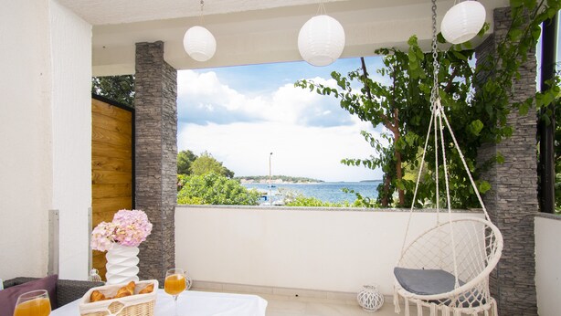 Un balcon avec une table blanche dressée pour le petit-déjeuner, un fauteuil suspendu et une vue sur la mer.