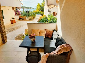 A balcony with a wicker sofa, a small table with a vase, and a view of a coastal area. - Apts Alisa- One bedroom apartment with terrace and sea view (Brodarica)