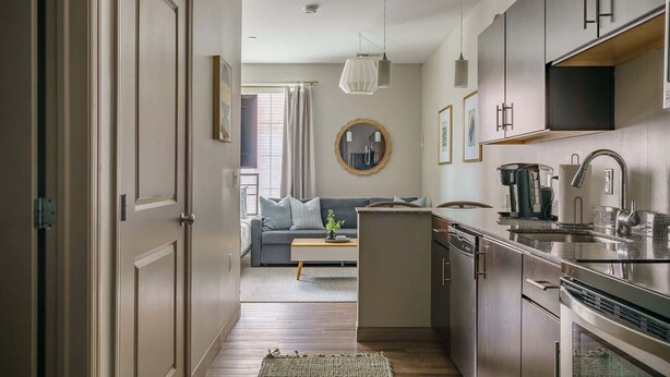 Una cocina moderna con electrodomésticos de acero inoxidable, un comedor con un sofá y una mesa de centro, y una sala de estar con un amplio ventanal y cortinas.
