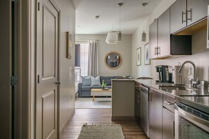 A modern kitchen with stainless steel appliances, a dining area with a sofa and a coffee table, and a living room with a large window and curtains.