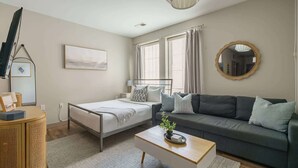 Un dormitorio moderno con cama, sofá, mesita de noche, lámpara, espejo y ventana con cortinas.