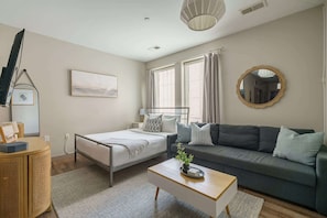 A modern bedroom with a bed, a sofa, a bedside table, a lamp, a mirror, and a window with curtains.