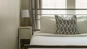 Una cama bien hecha con un cojín estampado, una lámpara de noche y una ventana con persianas.