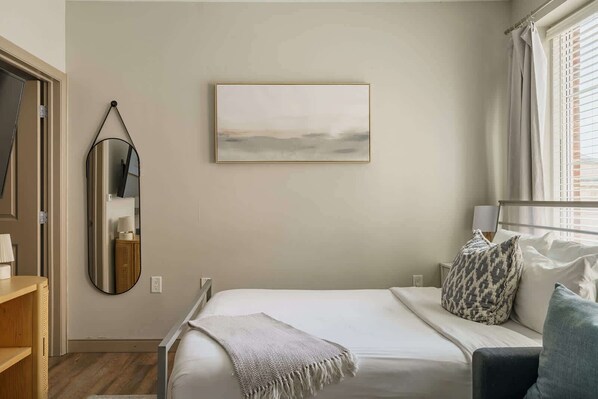A bedroom with a bed, a bedside table, a mirror, a wall-mounted picture, and a window with curtains.