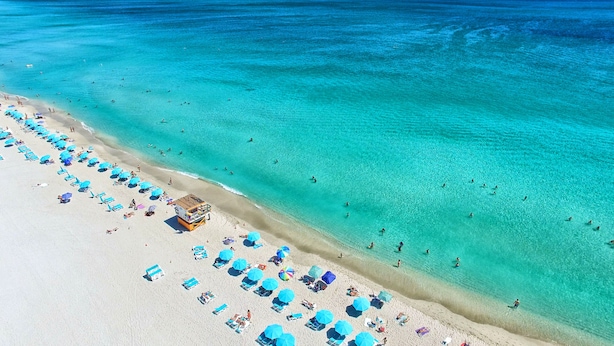 An aerial view of a beach with clear turquoise waters, numerous sun loungers, and umbrellas.