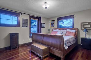 A bedroom with a wooden bed, a bench, a dresser, and two windows.